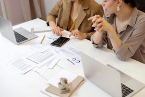 Two office workers talk while performing calculations and using laptops at a table